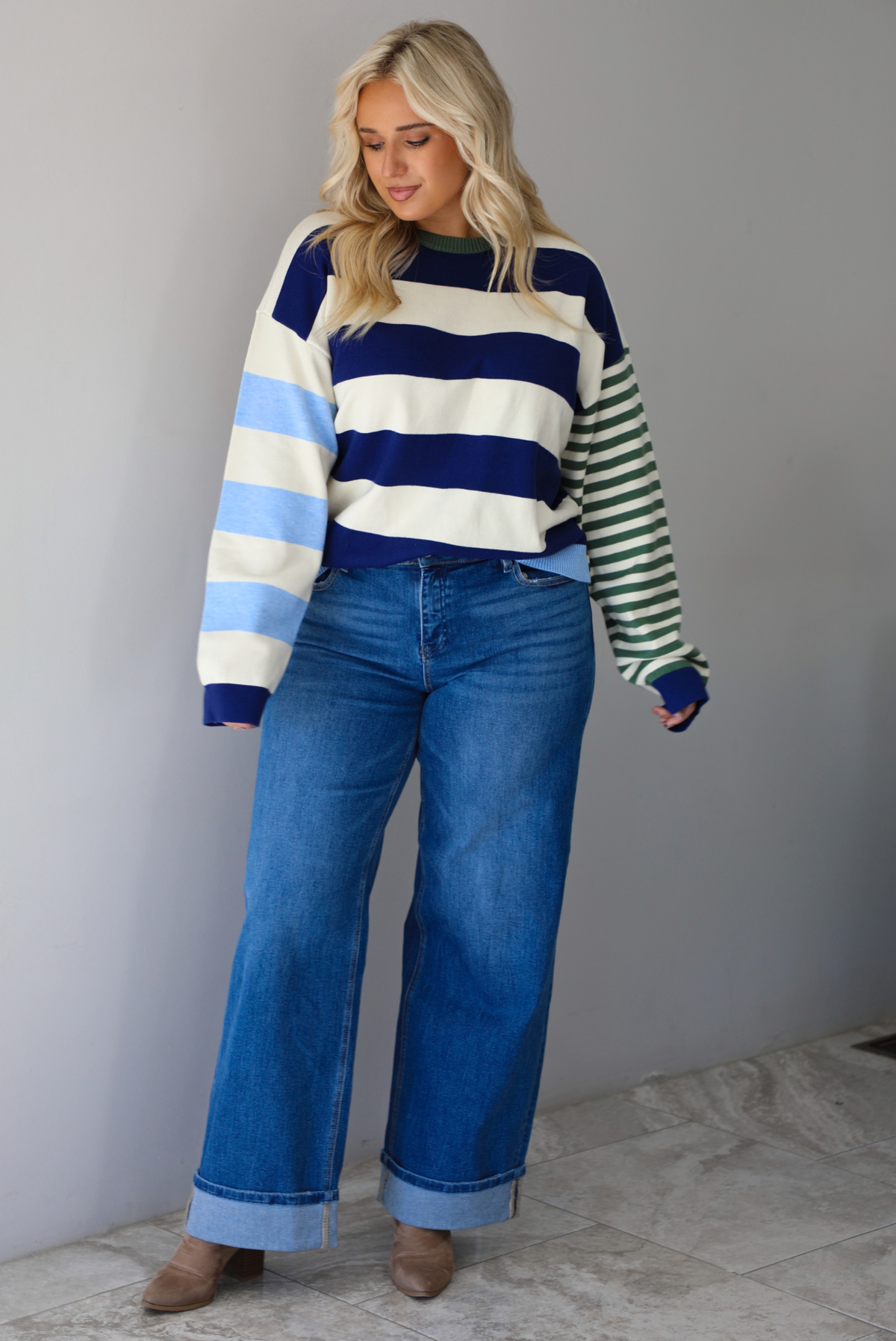 Woman wearing a navy, cream, blue, and green alternating striped design throughout, paired with dark denim jeans, standing indoors against a white wall with white marble flooring. 
