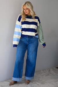 Woman wearing a navy, cream, blue, and green alternating striped design throughout, paired with dark denim jeans, standing indoors against a white wall with white marble flooring. 