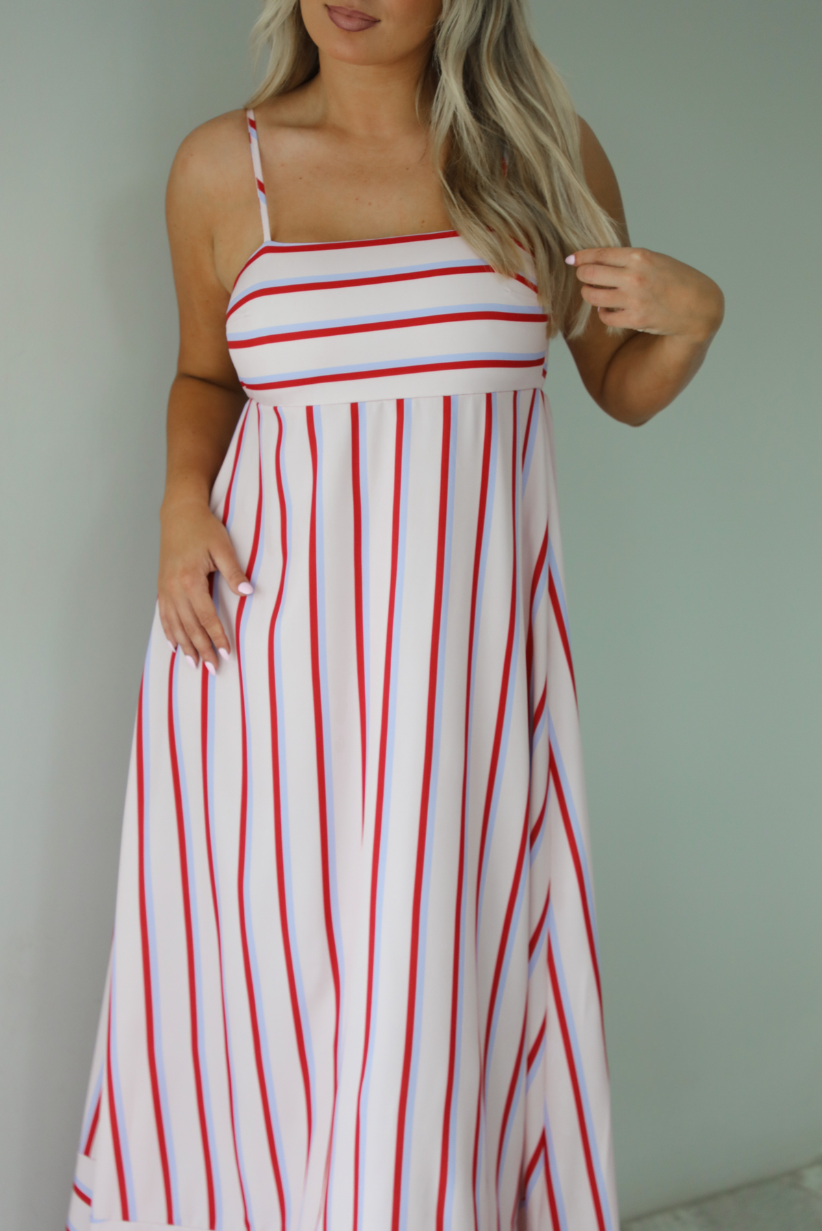 Woman wearing a spaghetti strap, maxi length dress that has a light pink color throughout with a red and light blue striped pattern. The photo is taken indoors against a white wall.