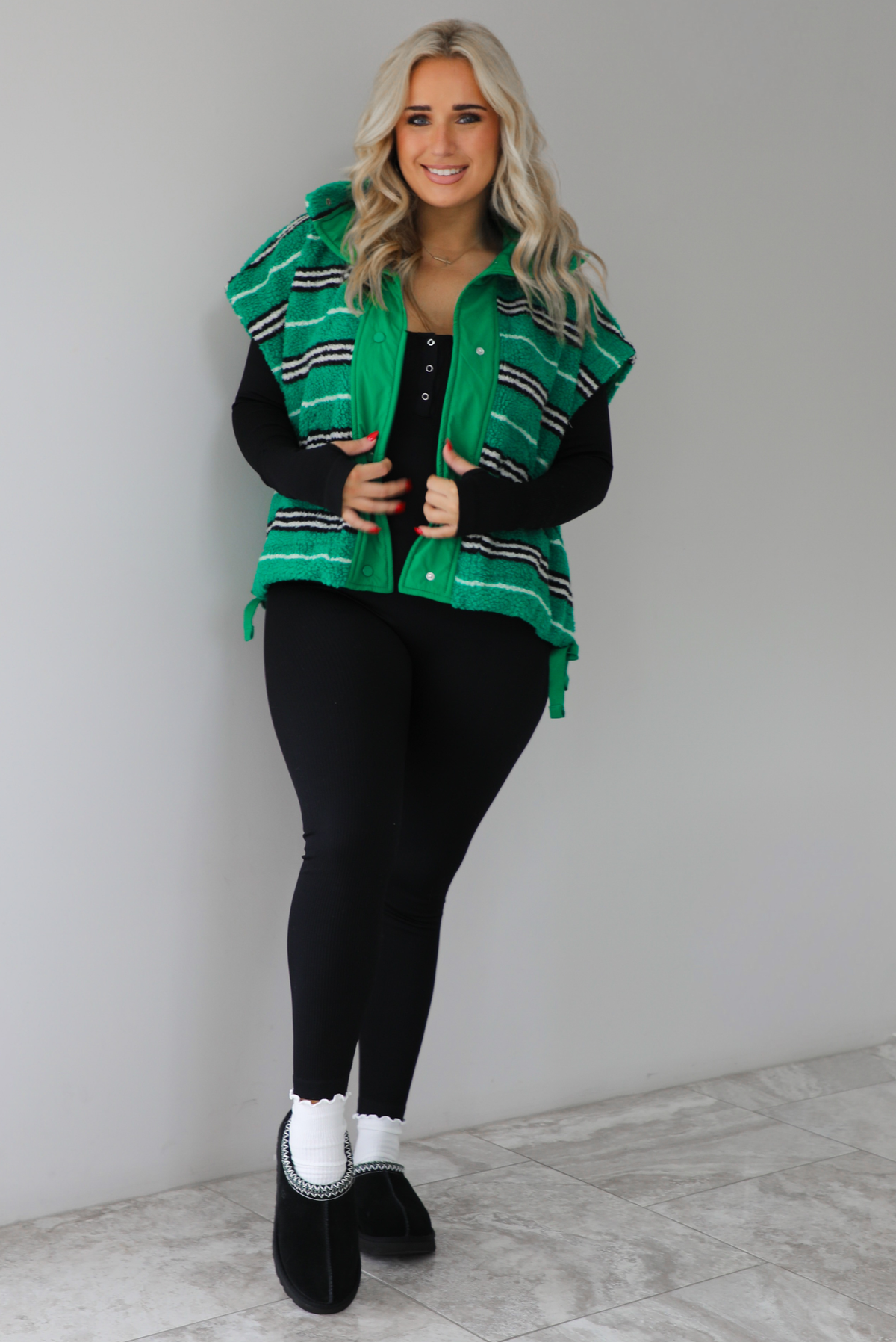 Woman wearing a green sherpa vest with black and white stripes that has an oversized fit, with a long sleeved, fitted, full body jumpsuit, with white slouchy socks and black ugg boot slides. The photo is taken indoors against a white wall with white marbled flooring. 