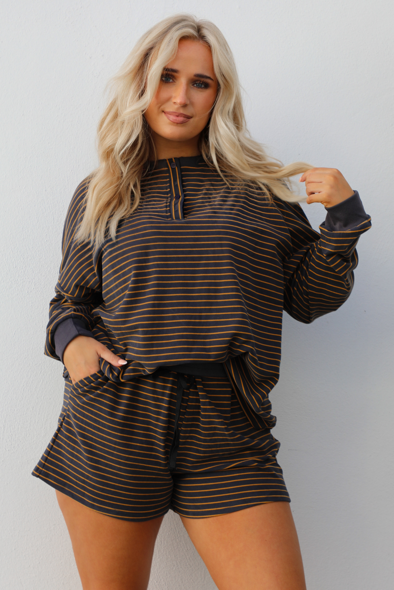 Close up of a woman model wearing a brown and multi-colored striped pattern throughout. The set consists of an oversized top with quarter-button details and shorts. The photo is taken outdoors against a white wall. 