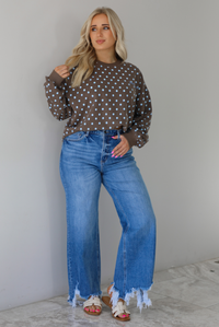 Woman wearing a brown sweater that has a blue polka dot print pattern throughout with blue embroidered writing on the left side that reads 'Trust The Process.' The sweater is paired with a medium wash denim. The photo is taken indoors against a white wall with white marble flooring.