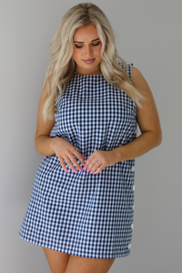 Woman wearing a navy and white gingham print patterned dress that has non-functional button closures on the left side. The photo is taken indoors against a white wall.
