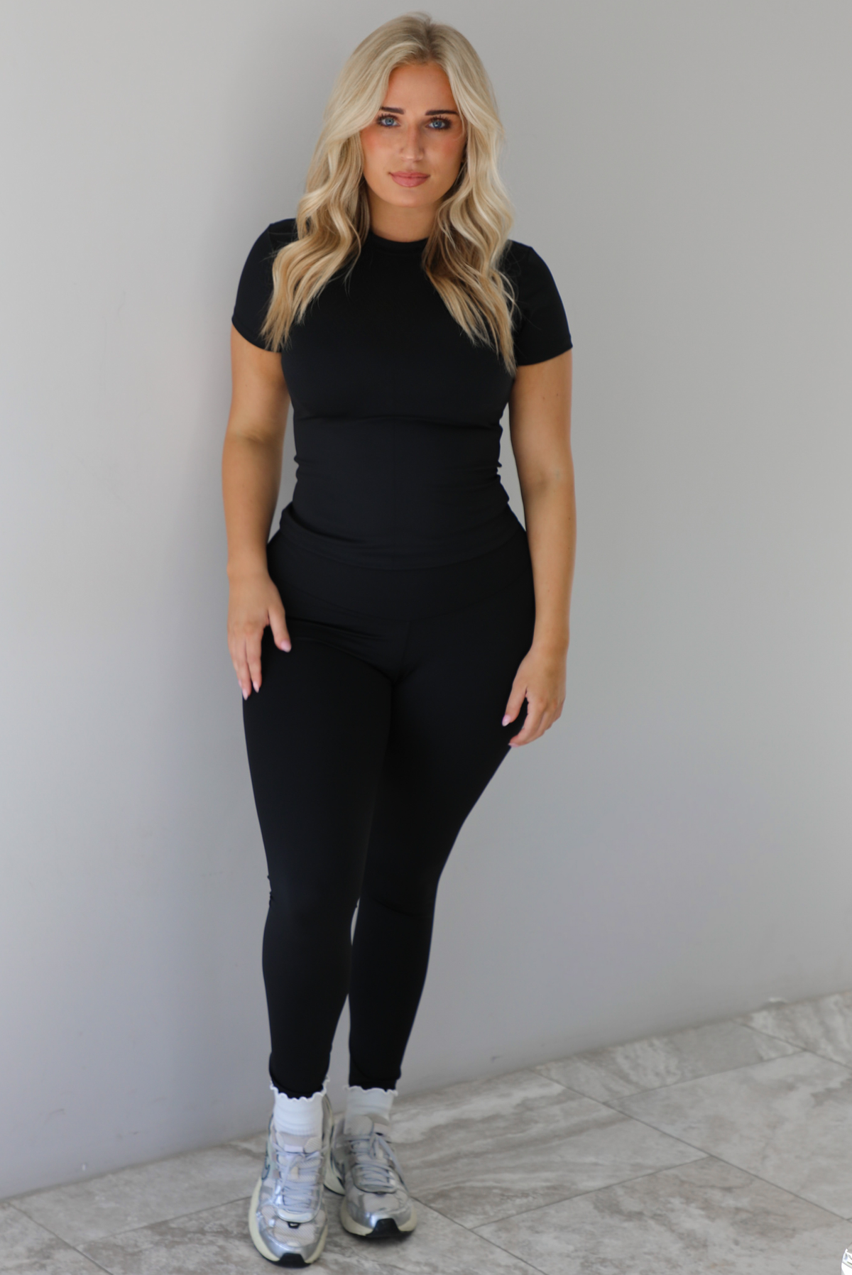 Woman wearing a fitted two piece black set featuring a short sleeved top and fitted leggings. The set is styled with a pair of white ankle socks and silver sneakers. The photo is taken indoors against a white wall with white marble flooring. 