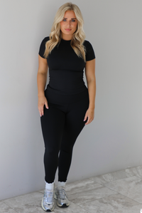 Woman wearing a fitted two piece black set featuring a short sleeved top and fitted leggings. The set is styled with a pair of white ankle socks and silver sneakers. The photo is taken indoors against a white wall with white marble flooring. 