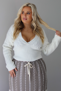 A woman wearing an ivory v-neck sweater paired with a taupe and white embroidered maxi skirt, standing indoors against a white wall.