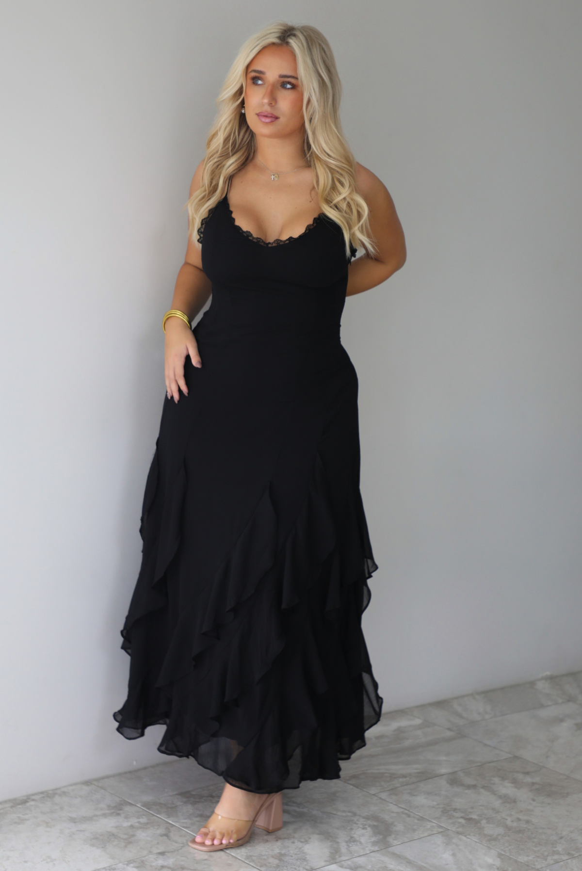 A woman wearing a black formal maxi dress with lace trim detailing at the bodice and ruffled details at the skirt, standing indoors against a white wall with white marble flooring. 