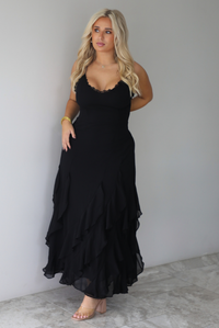 A woman wearing a black formal maxi dress with lace trim detailing at the bodice and ruffled details at the skirt, standing indoors against a white wall with white marble flooring. 