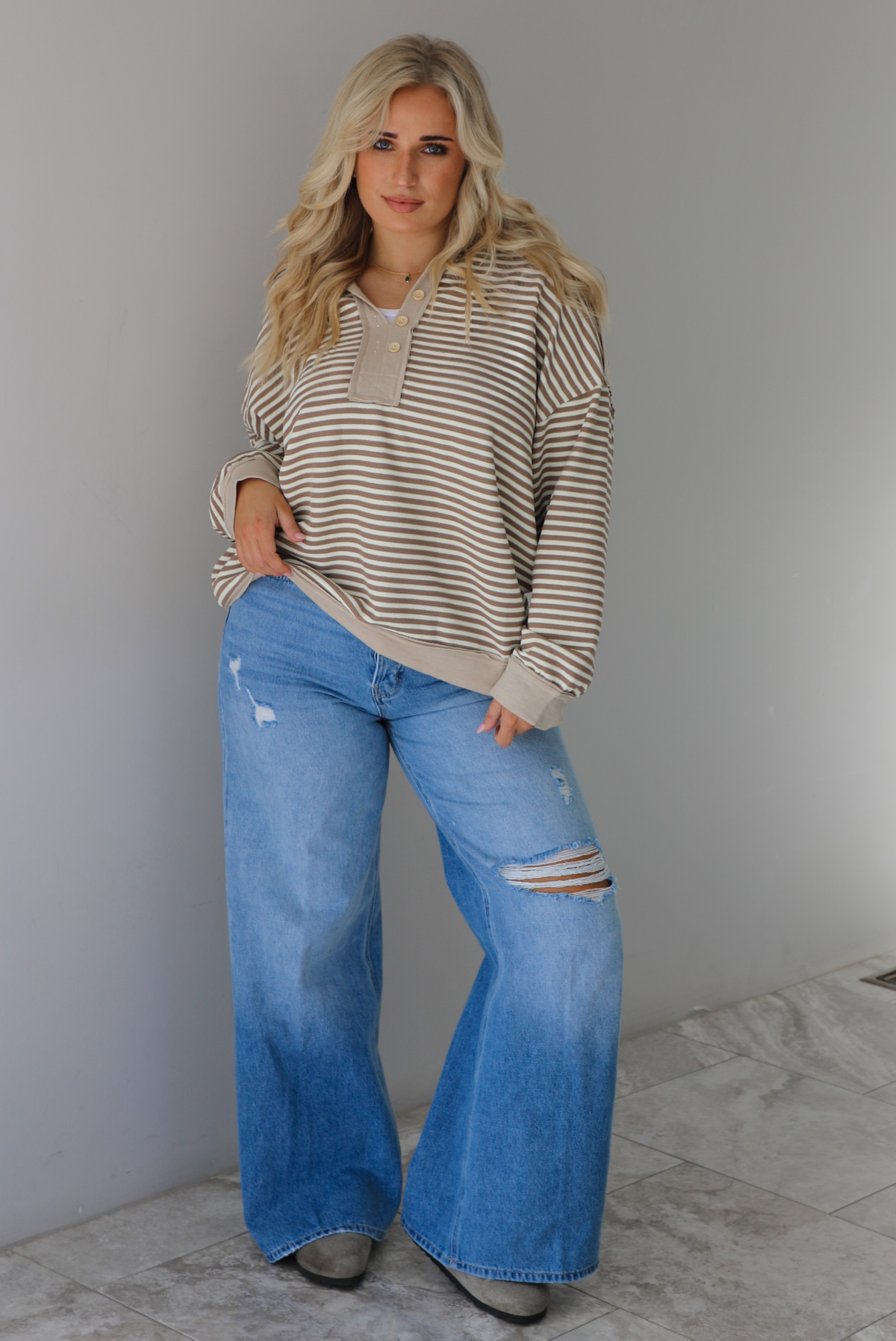 Woman wearing a mocha and cream striped pullover with a collared neckline, quarter button closures in the front, that has an oversized fit. The pullover is paired with a medium wash denim. The photo is taken indoors against a white wall with white marble flooring. 