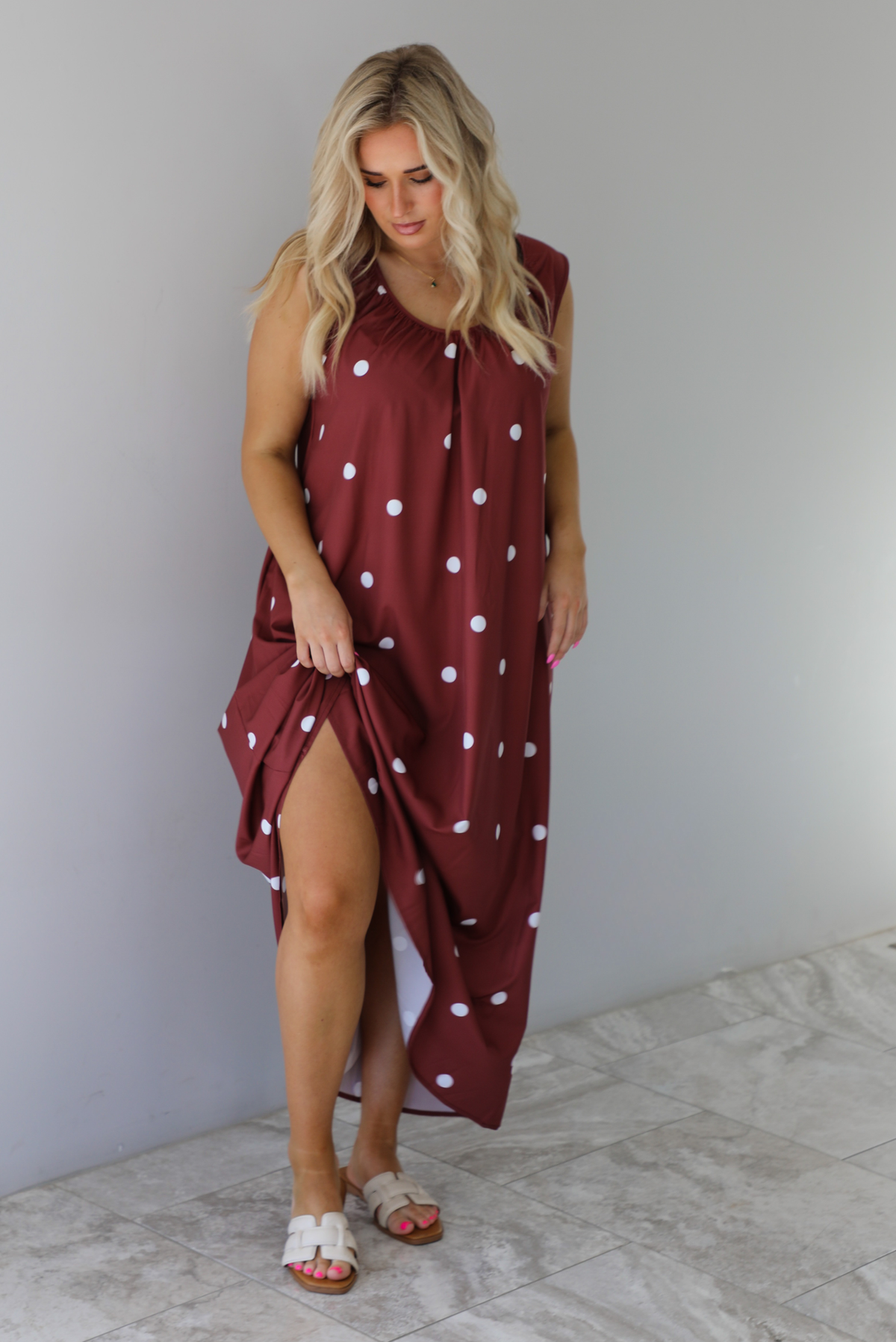 Woman wearing a red brown maxi length dress that has a white polka dot print pattern throughout. The dress is paired with sand sandals. The photo is taken indoors against a white wall with white marble flooring. 