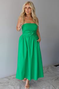 Woman wearing a green strapless midi dress that has a fitted bodice, belted waistline detail, functional pockets on either side, and a midi length indoors against a white wall with white marble flooring. 