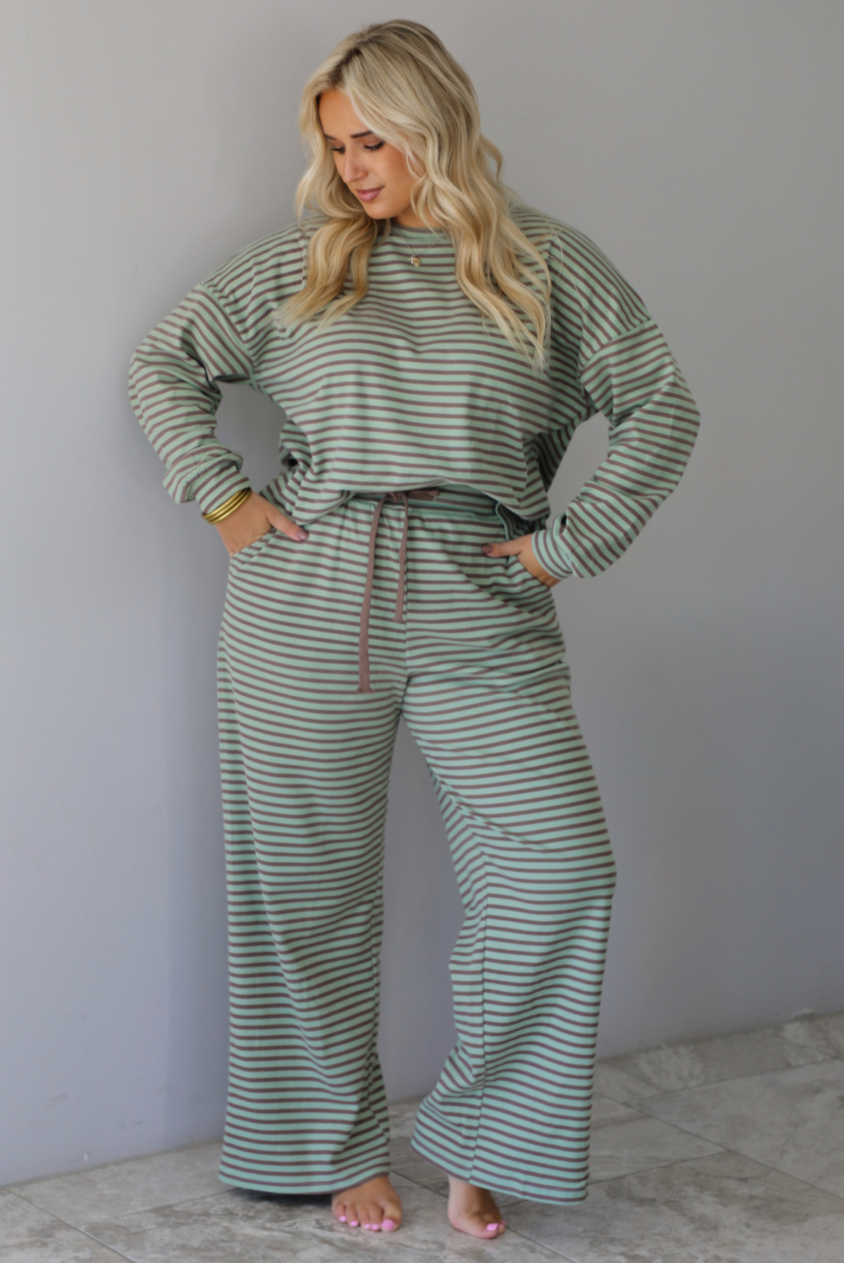 A woman wearing a pale green and brown striped two piece set featuring an oversized long sleeved top and high waisted, wide leg pants, standing indoors against a white wall with white marble flooring. 