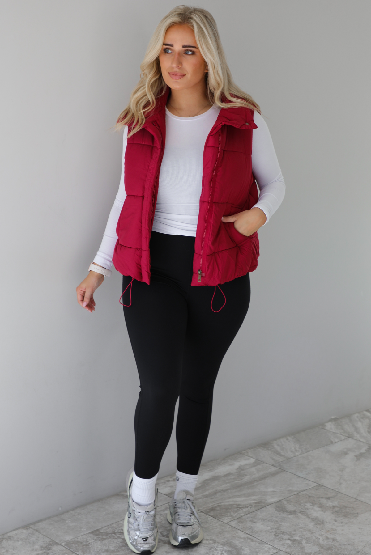Woman wearing a wine red colored puffer vest, white long-sleeve shirt, black leggings, and gray sneakers with white ankle socks indoors against a white wall with white marble flooring.