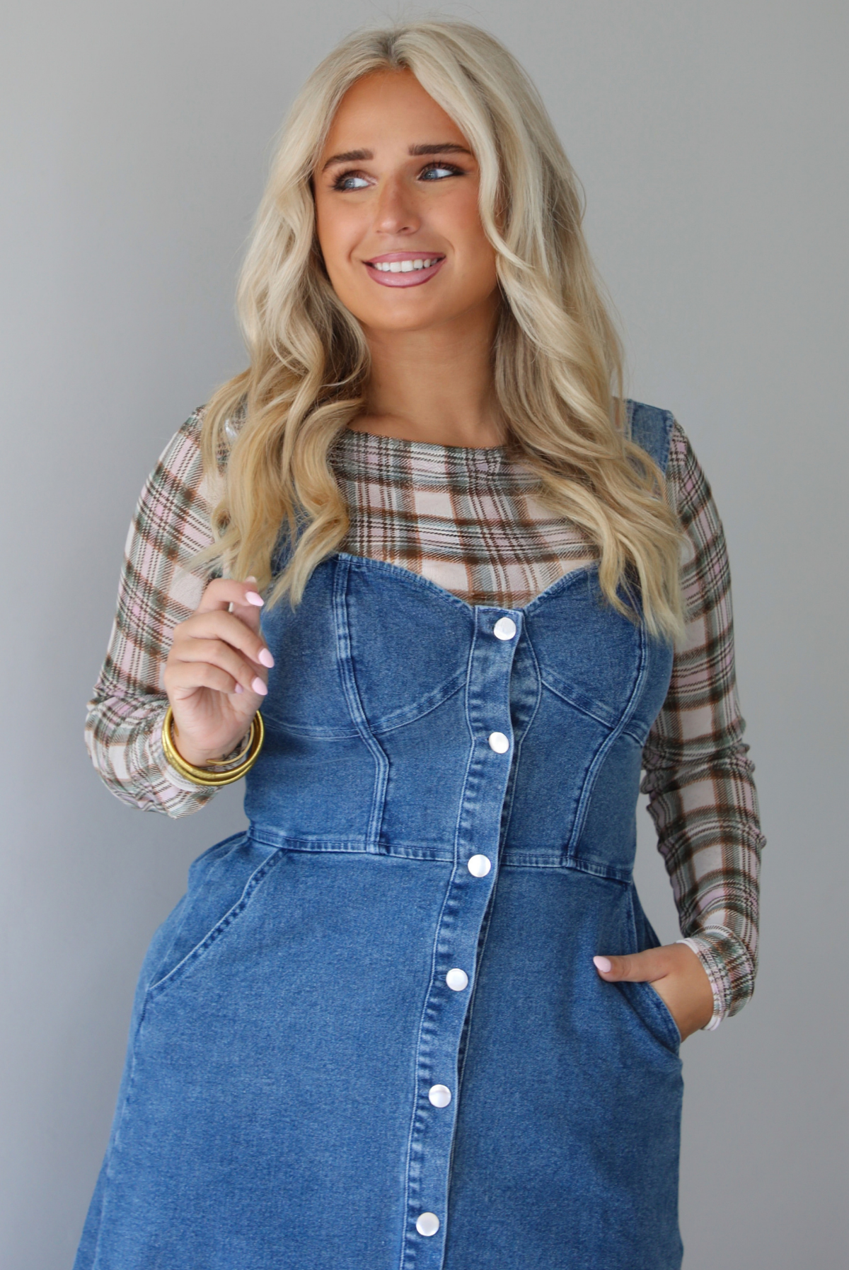 A woman wearing a medium wash denim midi dress with button closures along the  front, with pockets on either side, paired with a sheer, multi-colored plaid long sleeved top underneath, standing indoors against a white wall. The photo shows a close-up of the bodice, which has structured details that look similar to a corset top.
