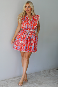 A woman modeling a sleeveless mini dress with a red, cream, and light blue paisley pattern, featuring a tie waist and ruffled collar, standing indoors against a white wall with white marble flooring. 