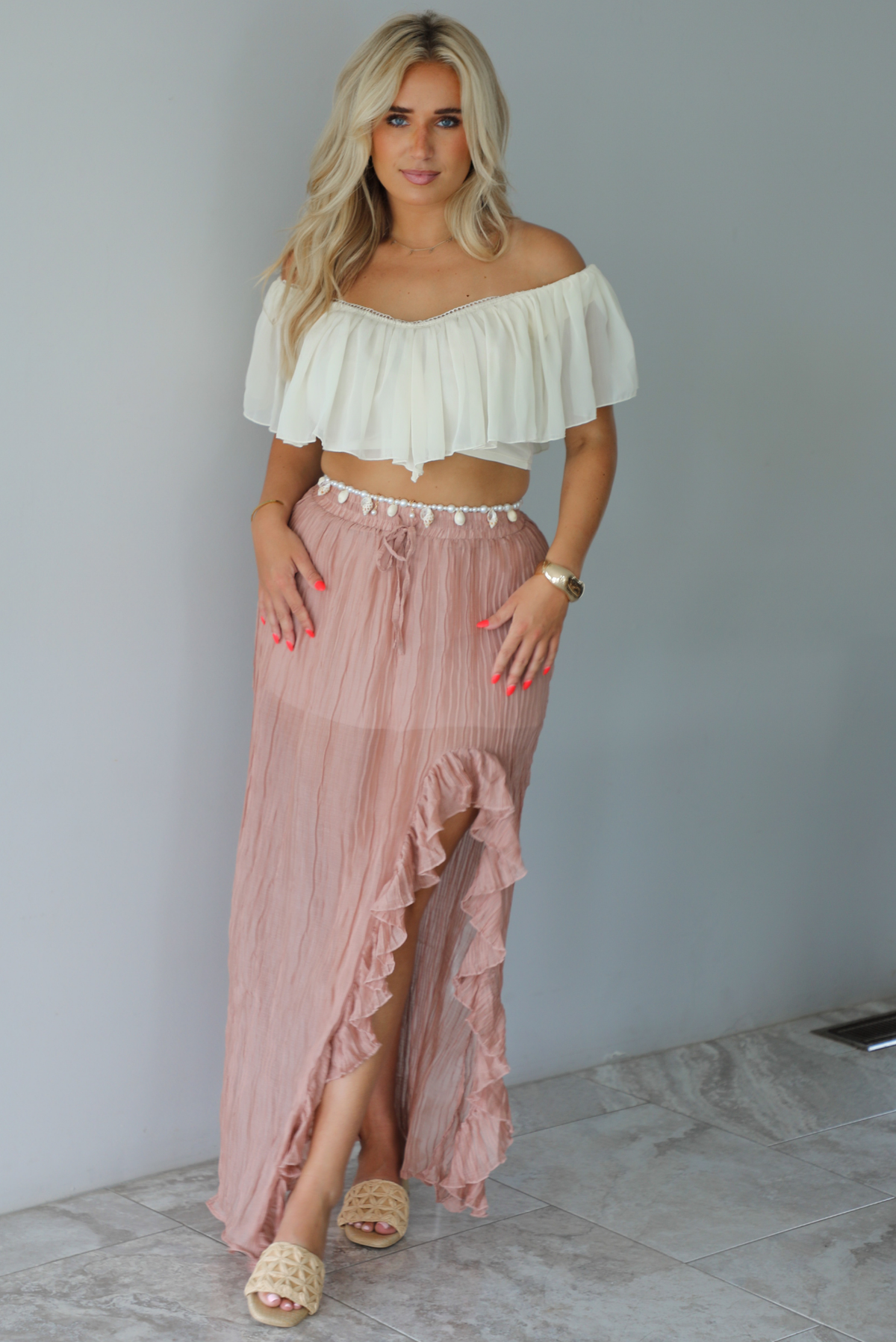 Woman wearing a cream cropped, off the shoulder top with a high waisted blush maxi skirt indoors against a white wall with white marble flooring. 