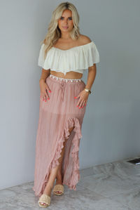 Woman wearing a cream cropped, off the shoulder top with a high waisted blush maxi skirt indoors against a white wall with white marble flooring. 