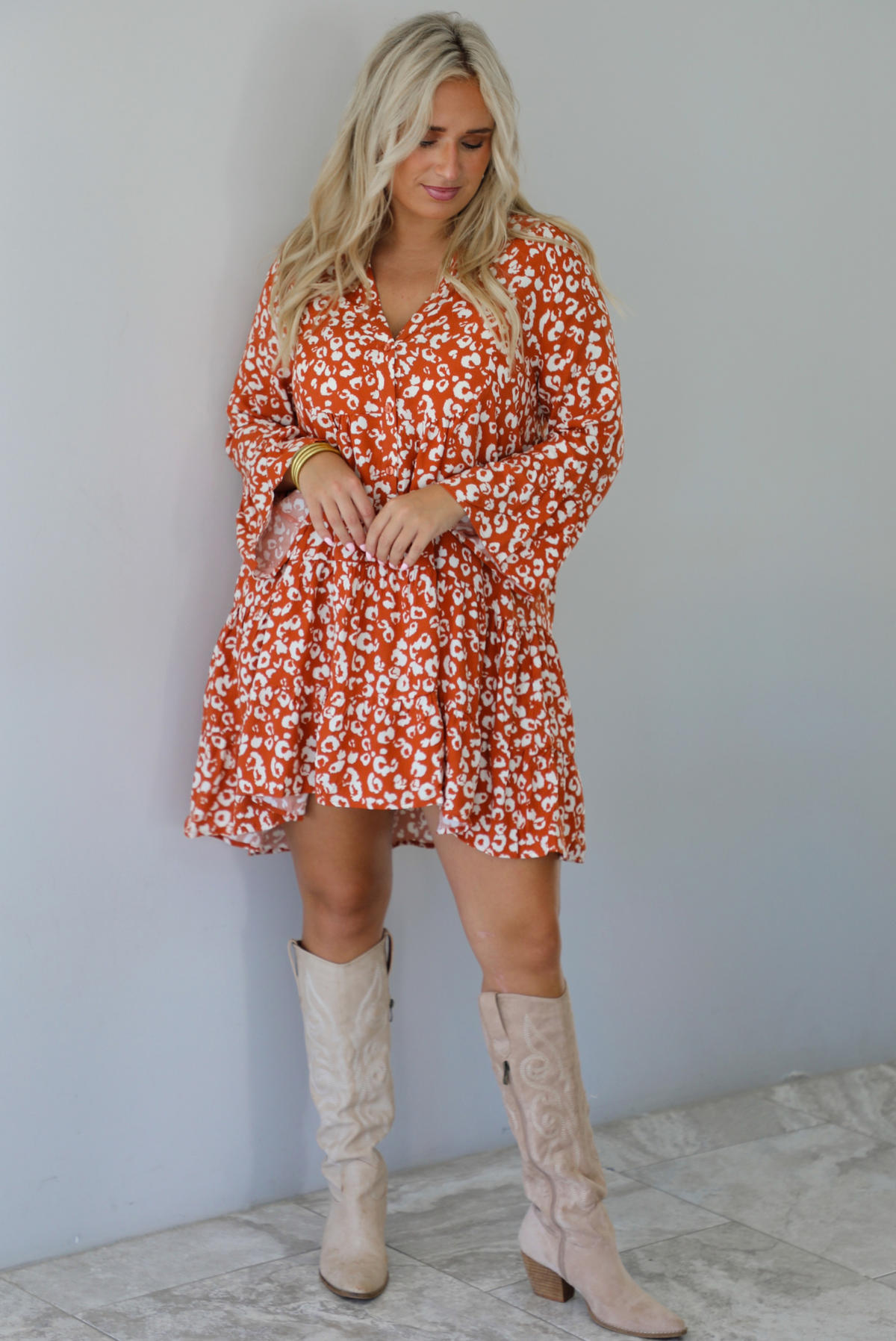 A woman wearing an orange and white animal print long sleeved, flowy mini dress, standing indoors against a white wall with white marble flooring. 