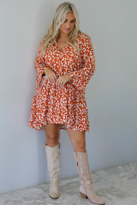A woman wearing an orange and white animal print long sleeved, flowy mini dress, standing indoors against a white wall with white marble flooring. 