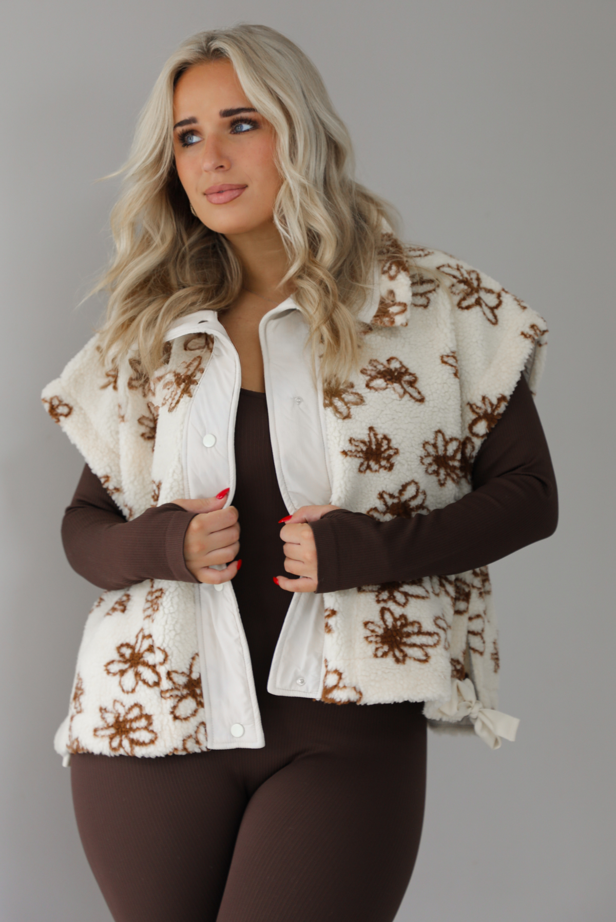 The photo features a model wearing a beige sherpa vest with a brown floral print pattern throughout, functional button closures in the front, and that is oversized with a brown, fitted jumpsuit. The photo is taken indoors against a white wall with white marbled flooring. 