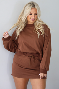 A woman wearing a brown matching two piece set featuring a cropped sweatshirt and mini skirt of the same material, standing indoors against a white wall. The model tucked the front of the top into the front of the skirt. 