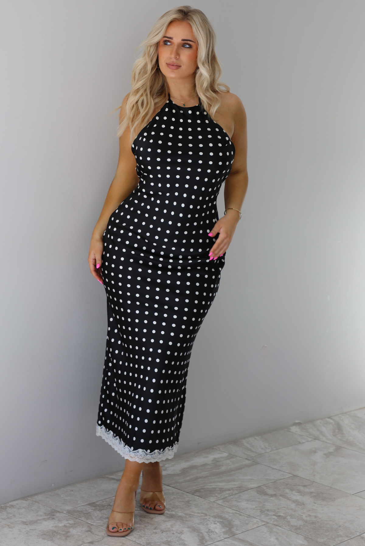Woman wearing a black wearing a white polka dot printed midi dress that has a halter tie neckline, is form fitting, with white lace trim at the bottom. The photo is taken indoors against a white wall with white marble flooring. 