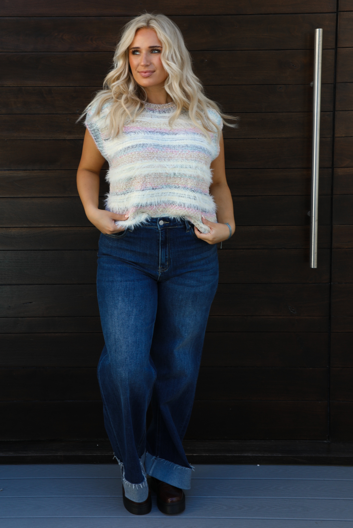 Woman model wearing a soft, striped sweater vest with dark wash denim jeans. The photo is taken outdoors against a dark brown wooden door. 