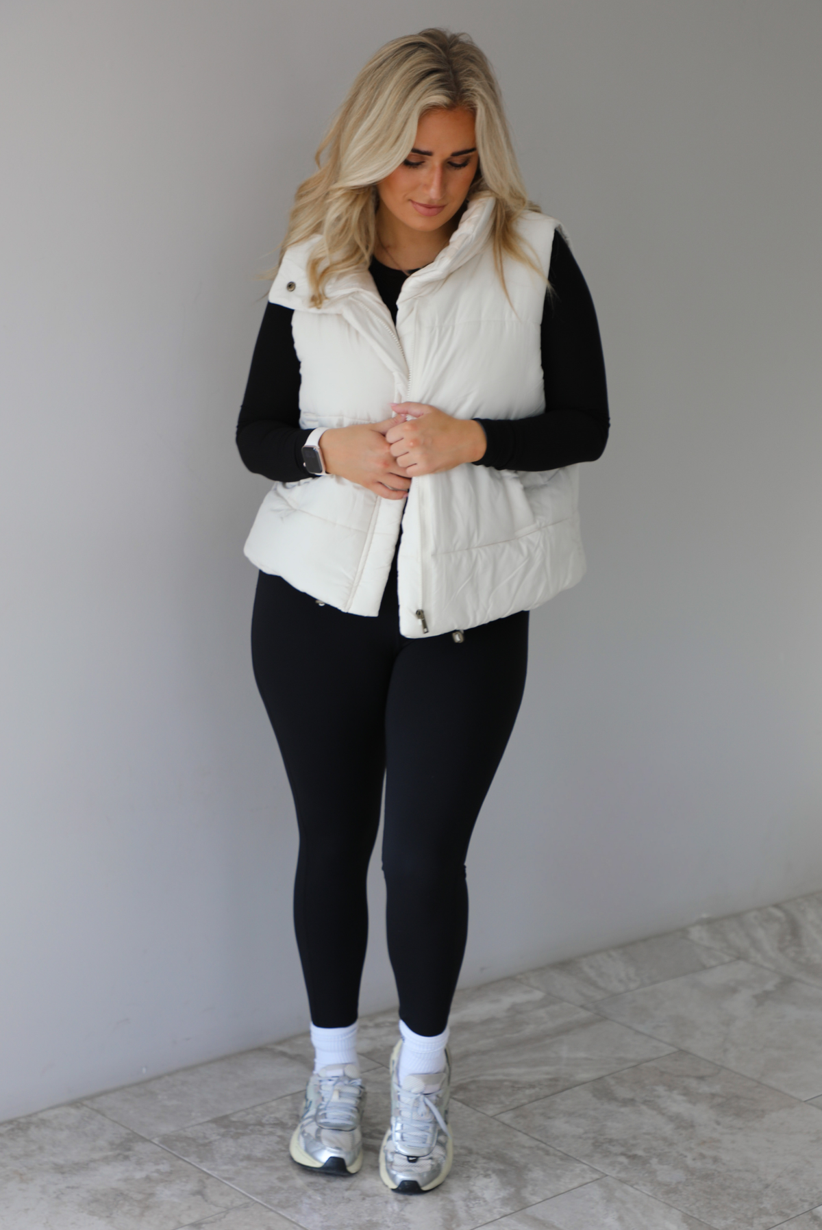 Woman wearing a light sand colored puffer vest with a fitted black long sleeved top and fitted leggings with high white ankle socks and silver sneakers. The photo is taken indoors against a white wall with white marble flooring. 