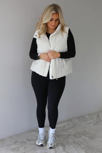Woman wearing a light sand colored puffer vest with a fitted black long sleeved top and fitted leggings with high white ankle socks and silver sneakers. The photo is taken indoors against a white wall with white marble flooring. 