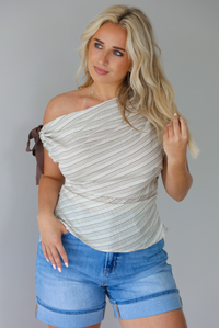 Woman wearing a multi-colored striped top that has an off the shoulder design with a tie on the right side with medium wash denim shorts indoors against a white wall.