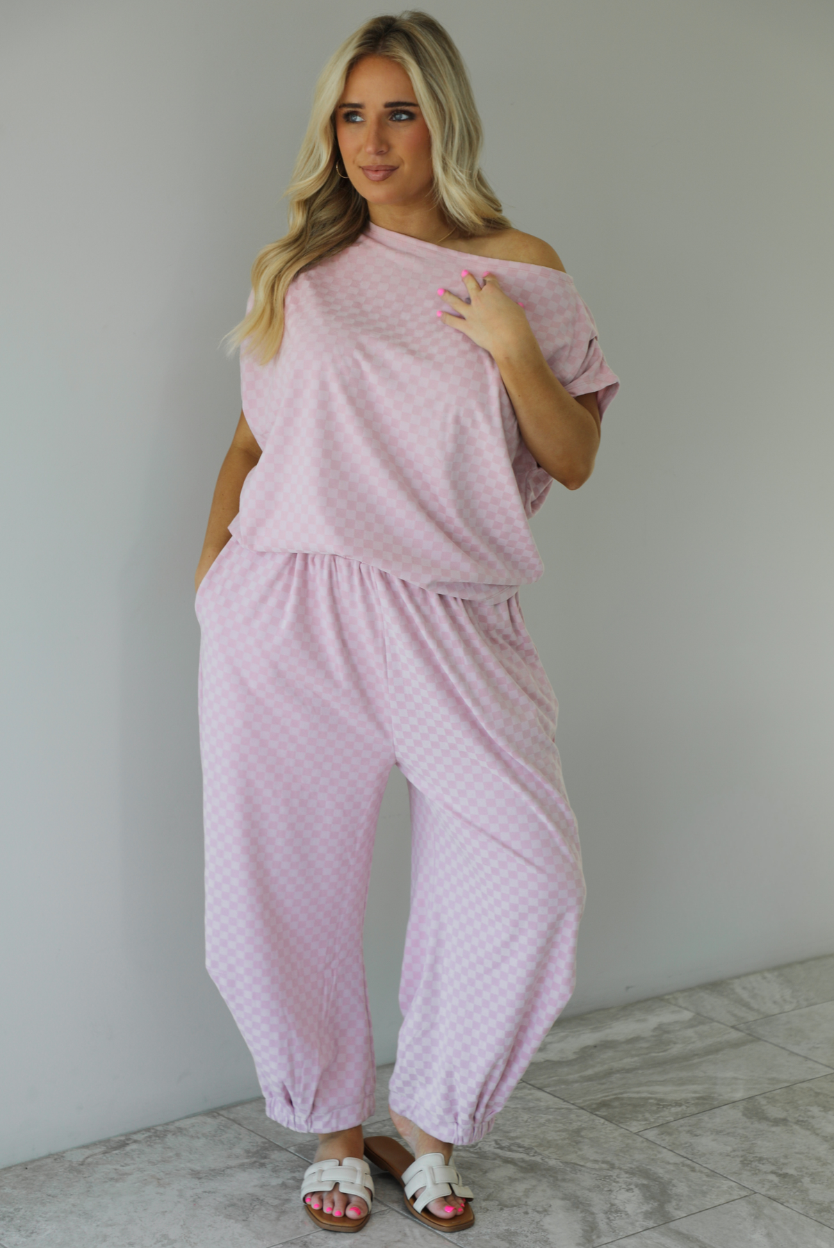 Woman wearing a pink checkered set featuring a slouchy top and pants. The photo is taken indoors against a white wall with white marble flooring.