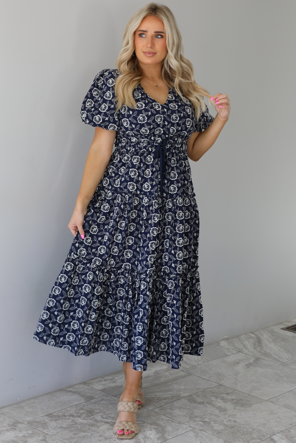 Woman wearing a navy dress that has a white embroidered floral print pattern throughout, a cinched waistline, and flowy maxi length skirt. The photo is taken indoors against a white wall with white marble flooring. 