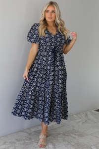 Woman wearing a navy dress that has a white embroidered floral print pattern throughout, a cinched waistline, and flowy maxi length skirt. The photo is taken indoors against a white wall with white marble flooring. 