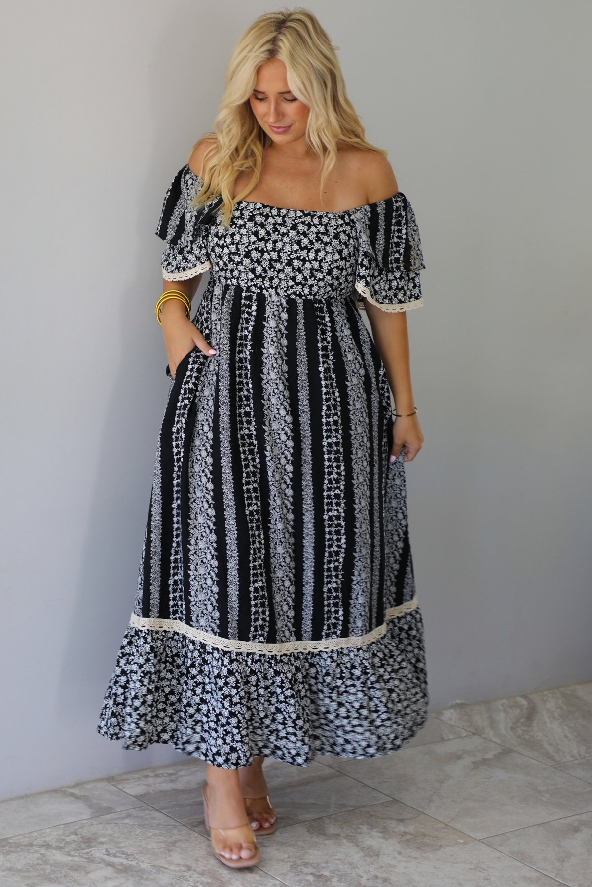 A woman wearing a black and white maxi dress with a white floral print that alternates between a cluster appearance and a striped appearance, with off the shoulder sleeves and a flowy skirt, standing indoors against a white wall with white marble flooring. 