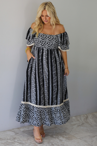 A woman wearing a black and white maxi dress with a white floral print that alternates between a cluster appearance and a striped appearance, with off the shoulder sleeves and a flowy skirt, standing indoors against a white wall with white marble flooring. 