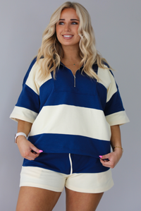 Woman wearing a navy and pale yellow thick striped set featuring a collared short sleeved quarter zip top and high waisted shorts that have an elastic drawstring waistline with functional pockets on either side. The photo shows the top untucked from the shorts. The photo is taken indoors against a white wall.