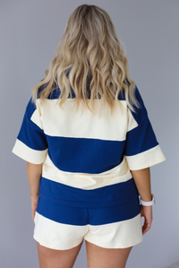 Woman wearing a navy and pale yellow thick striped set featuring a collared short sleeved quarter zip top and high waisted shorts that have an elastic drawstring waistline with functional pockets on either side. The photo shows the back of the set. The photo is taken indoors against a white wall.