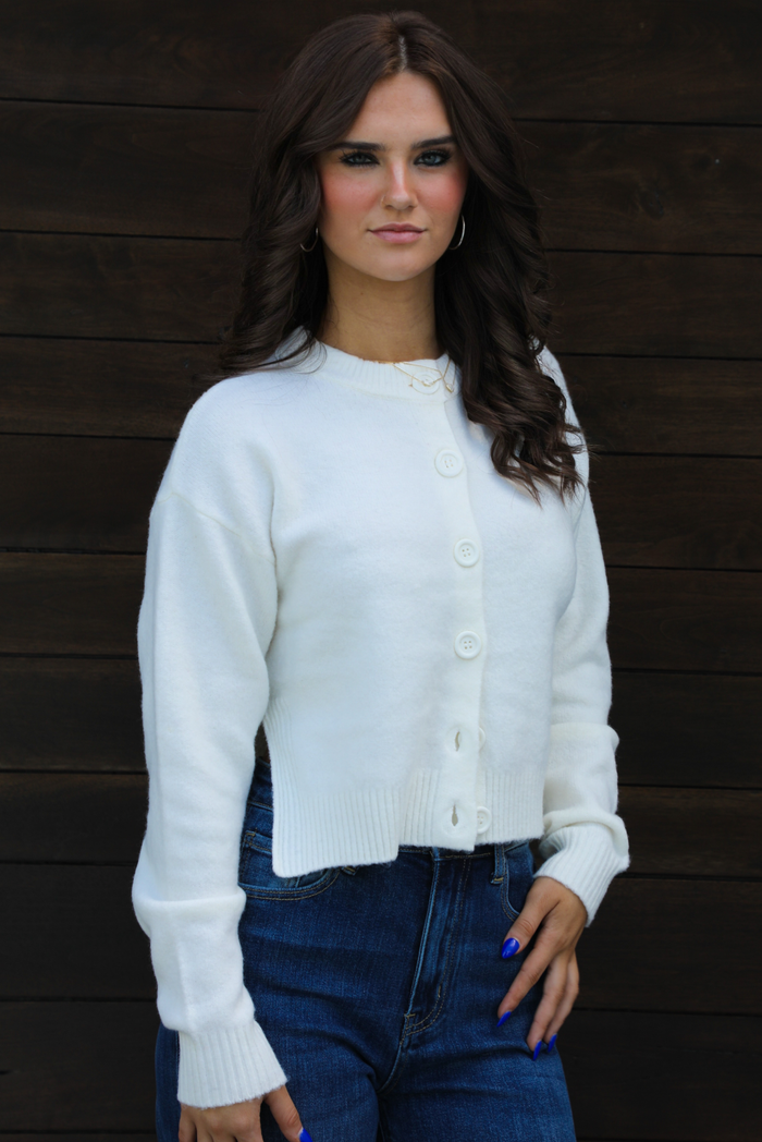 Woman model wearing an off-white cardigan that has a high neckline, functional button closures in the front, long sleeves, a slightly cropped cut, and split hem details on either side. The cardigan is paired with high waisted dark wash denim. The photo shows a close u p of the cardigan. The photo is taken outdoors against a dark brown wooden door.