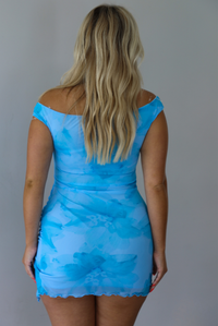 A woman viewed from behind wearing a bright blue off-the-shoulder bodycon mini dress with a watercolor-like pattern, set against a white wall.