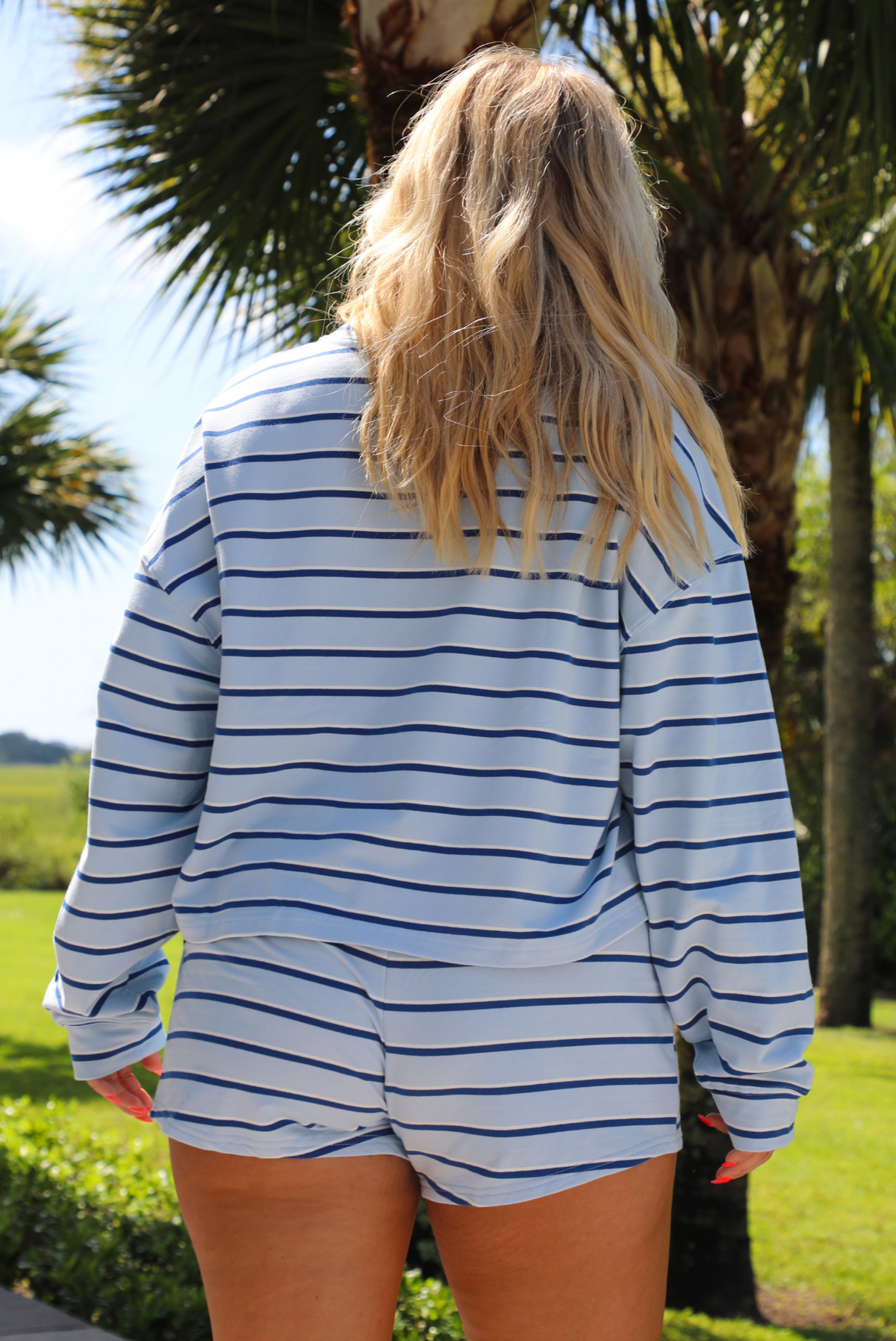 Woman wearing a blue, navy and white striped two piece set featuring a long sleeved tee and shorts outdoors with greenery and palm trees in the background.  The photo shows the back of the set. 