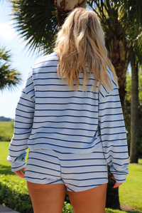 Woman wearing a blue, navy and white striped two piece set featuring a long sleeved tee and shorts outdoors with greenery and palm trees in the background.  The photo shows the back of the set. 