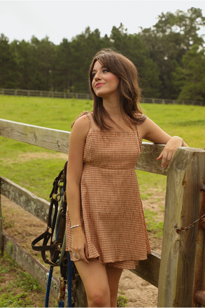 Woman wearing a peach and cream gingham patterned romper that looks like a dress. The photo is taken in a pasture, with the model leaning on a wooden fence, and bridles hanging behind her. The photo shows the adjustable tie straps. 