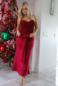 Woman model wearing a strapless, red holiday maxi length dress. The dress has a corset style bodice, a tie detail at the waistline, and a stretchy, satin textured maxi length skirt. The photo is taken inside a modern home in front of a decorated Christmas tree. 