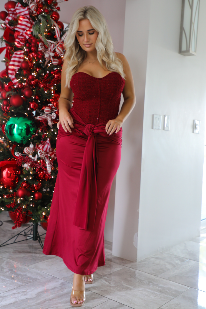 Woman model wearing a strapless, red holiday maxi length dress. The dress has a corset style bodice, a tie detail at the waistline, and a stretchy, satin textured maxi length skirt. The photo is taken inside a modern home in front of a decorated Christmas tree. 