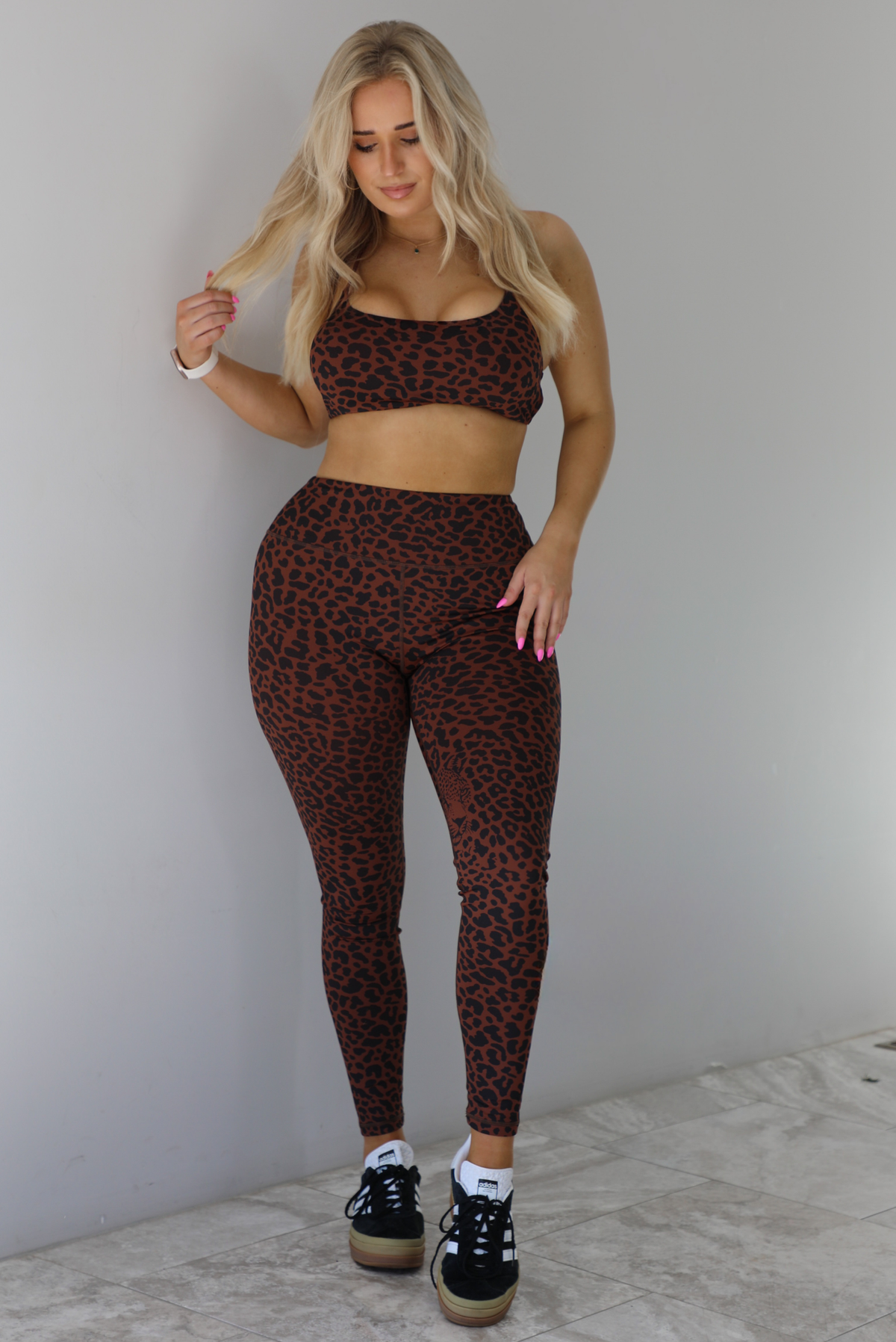 Woman wearing a brown and white leopard print two piece athleisure set that consists of a sports bra and high waisted leggings. The photo is taken indoors against a white wall with white marble flooring. 