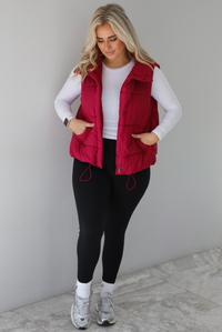 Woman wearing a wine red colored puffer vest, white long-sleeve shirt, black leggings, and gray sneakers with white ankle socks indoors against a white wall with white marble flooring.