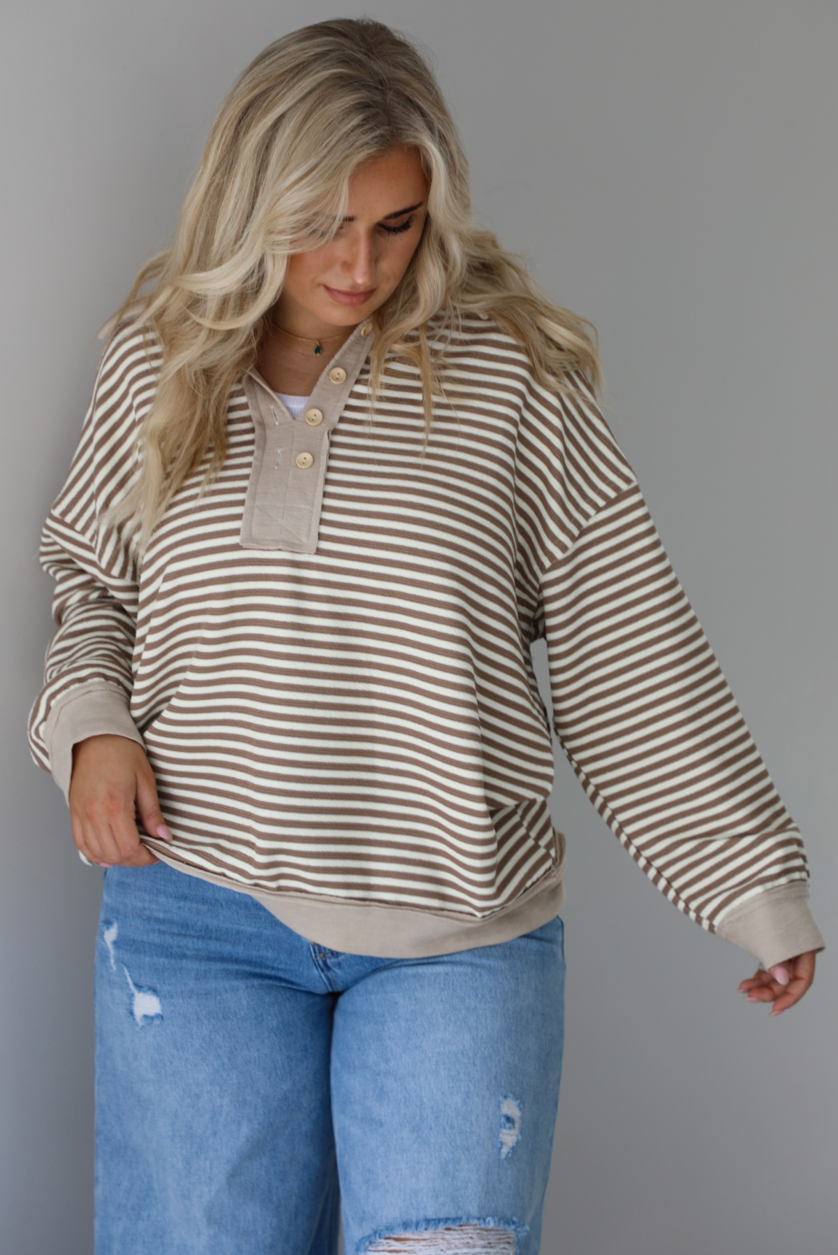 Woman wearing a mocha and cream striped pullover with a collared neckline, quarter button closures in the front, that has an oversized fit. The photo is taken indoors against a white wall.