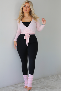Woman wearing a pink cropped wrap style fitted top over a black fitted jumpsuit. The outfit is paired with pink leg warmers. The photo is taken indoors against a white wall with white marble flooring. 