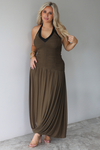 A woman wearing an olive maxi dress with black trim detailing at the v-halter neckline, has a fitted bodice and drop waist design, with a maxi-length flowy skirt, standing indoors against a white wall with white marble flooring. 