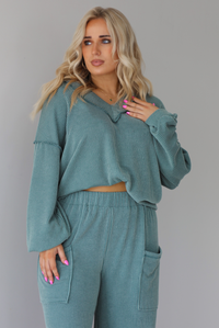 Woman wearing a teal two piece set that has a v-neck long sleeved oversized top and high waisted pants. The top is tucked into the woman's bra to give it a cropped look. The photo is taken indoors against a white wall.